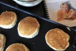 pancakes cooking on a griddle with batter and cookbook in background