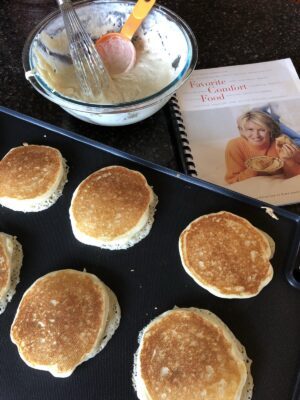 pancakes cooking on a griddle with batter and cookbook in background