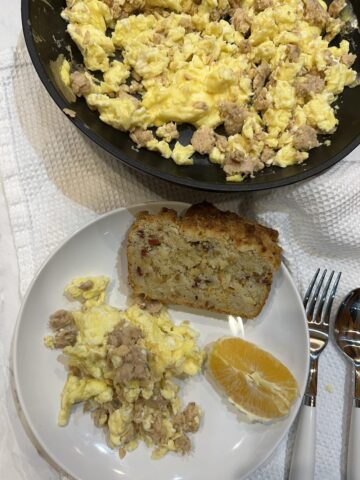eggs with salmon, bread, and an orange on a white plate with pan of eggs in background