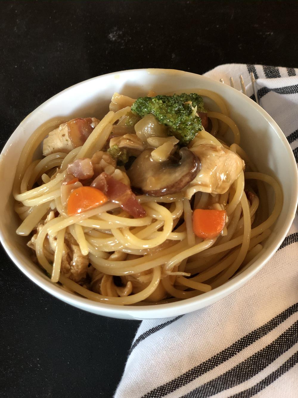 chicken bacon lo mein in white bowl on black and white napkin