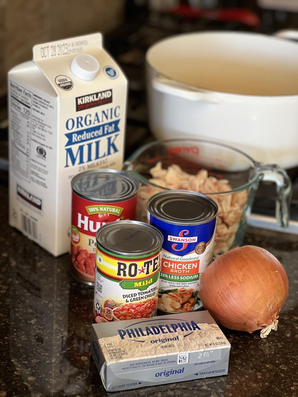 milk, chicken, canned tomatoes, chicken broth, onion and cream cheese on counter next to stove with a pan on it.