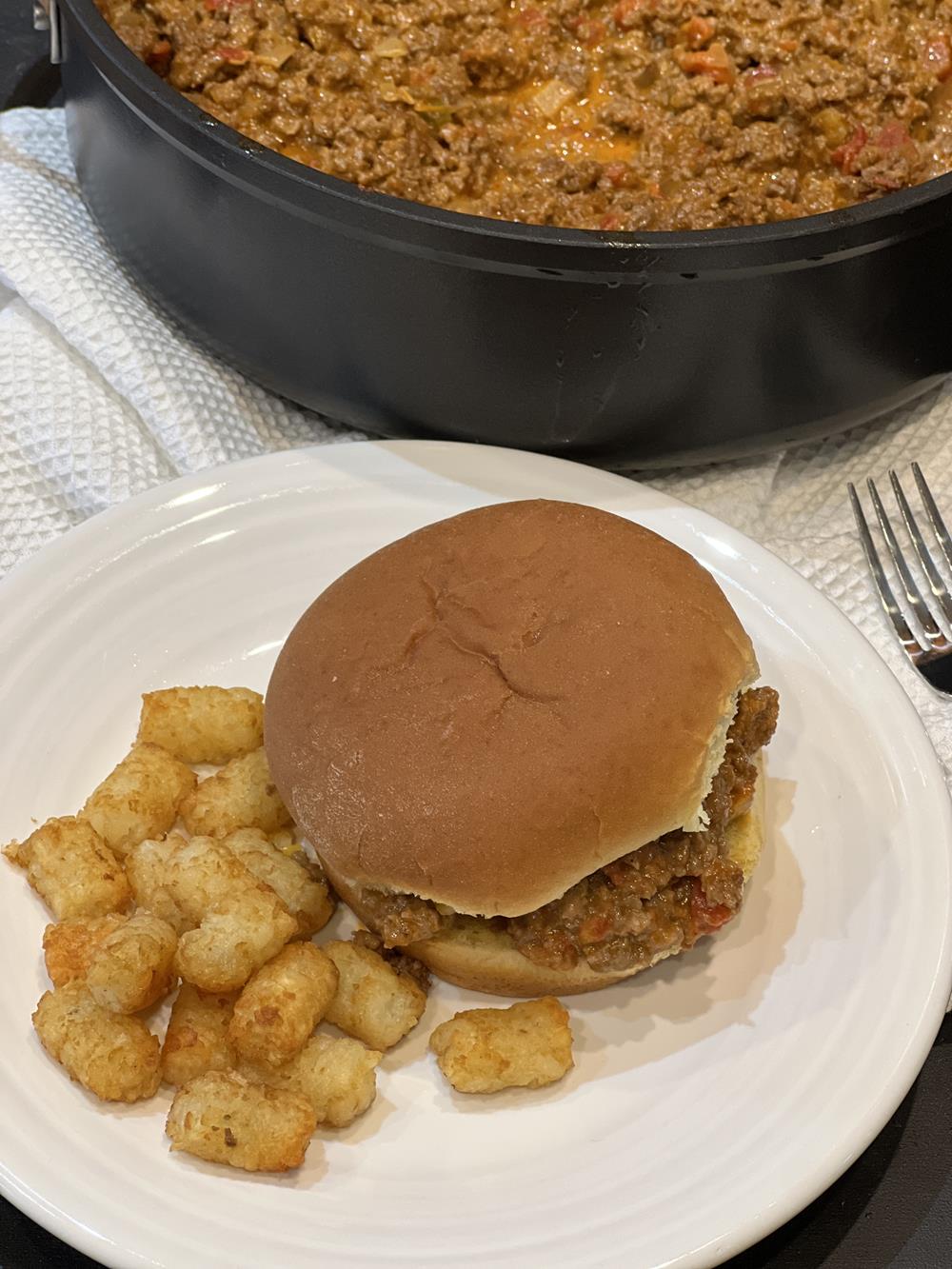 Sloppy joes on bun with tater tots on white plate with pan of sloppy joes in background