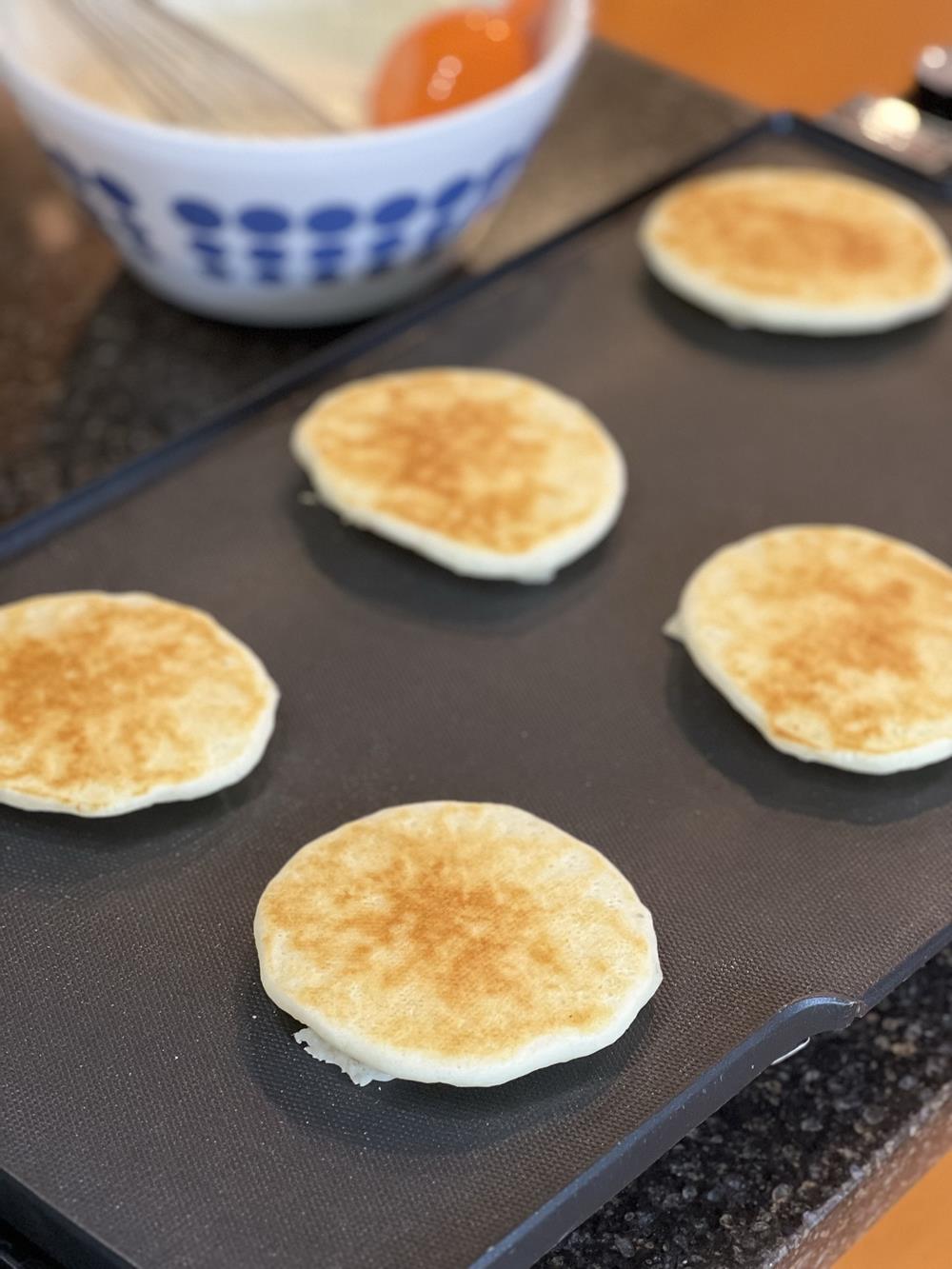 pancakes cooking on a griddle with bowl of batter in background