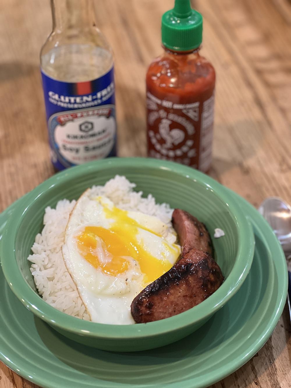 eggs and rice in green bowl with soy sauce and hot sauce in background