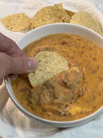 Velveeta chili in white bowl with hand dipping a tortilla chip in chili