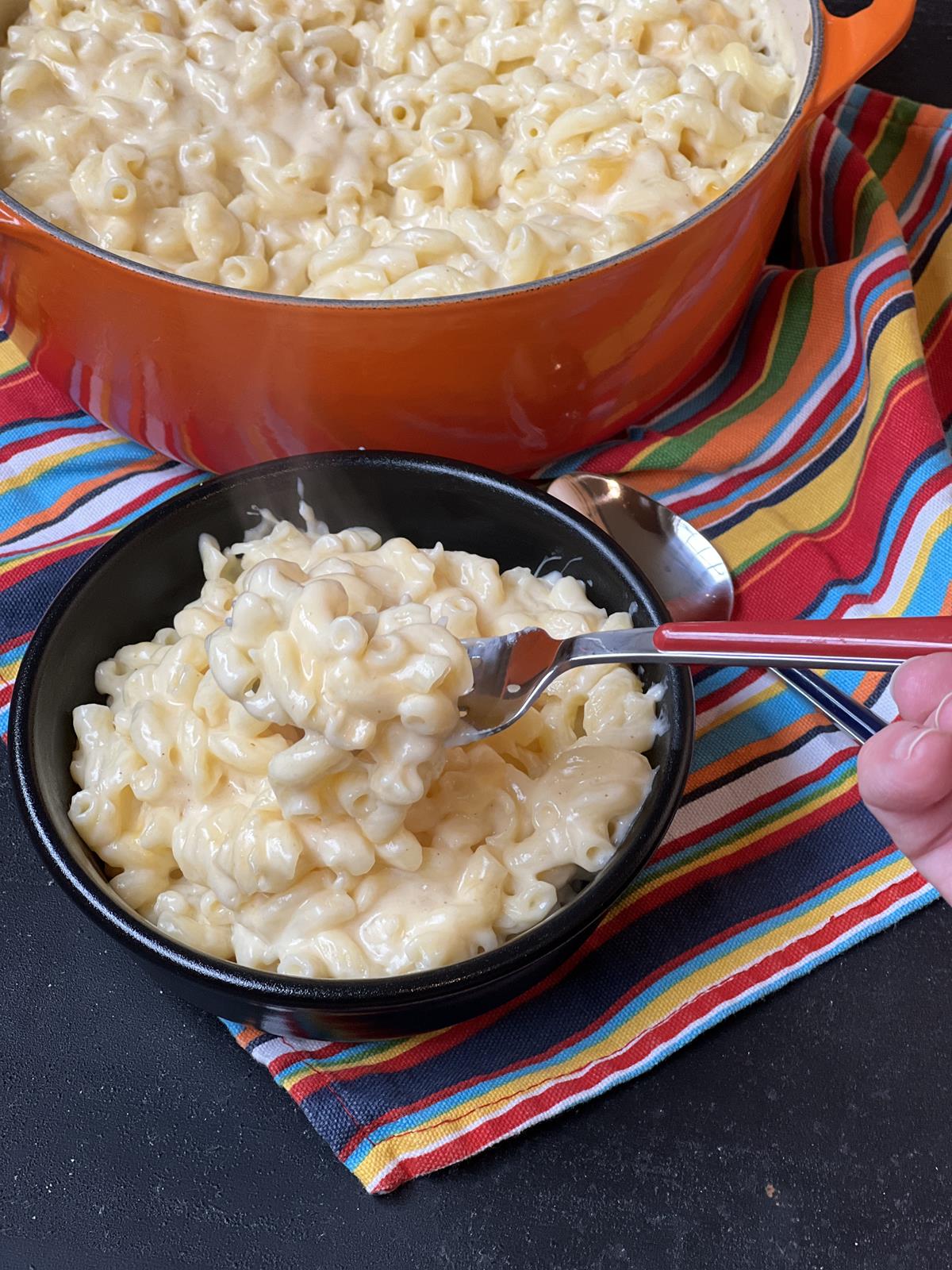 homemade mac and cheese in black bowl with red handled fork with pot of mac and cheese in background