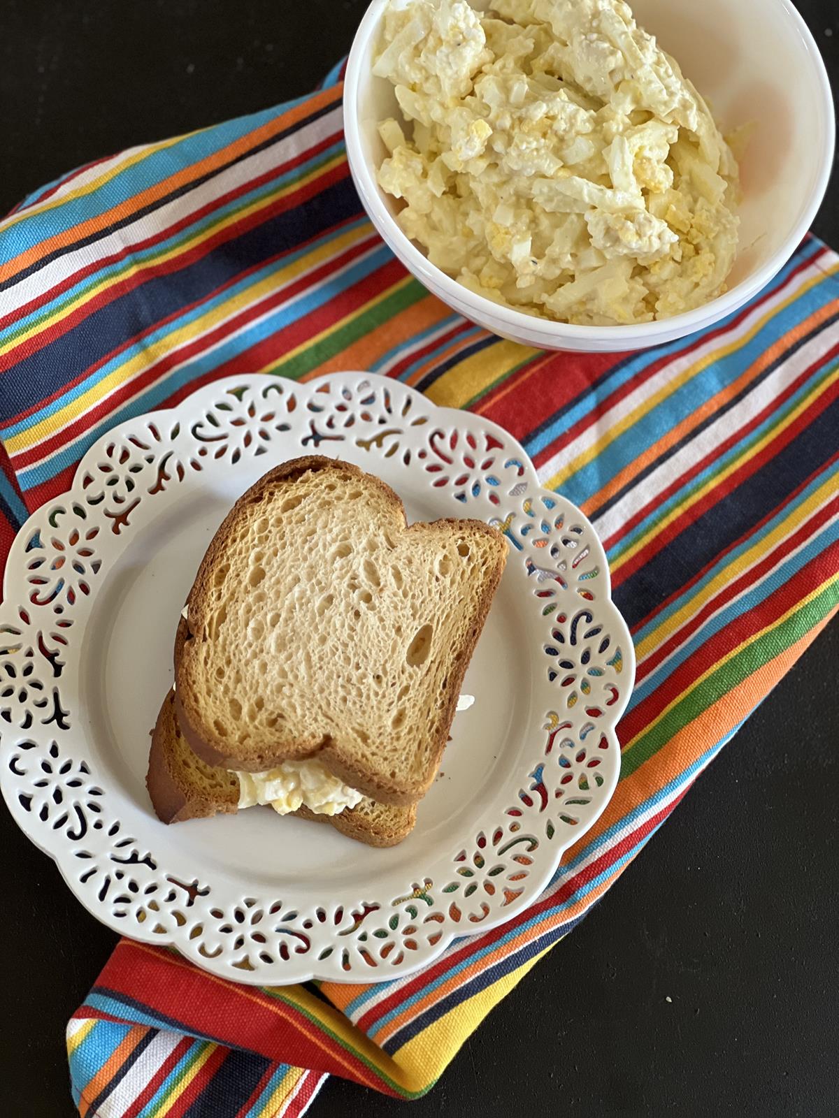egg salad sandwich on white plate with bowl of egg salad on a striped napkin