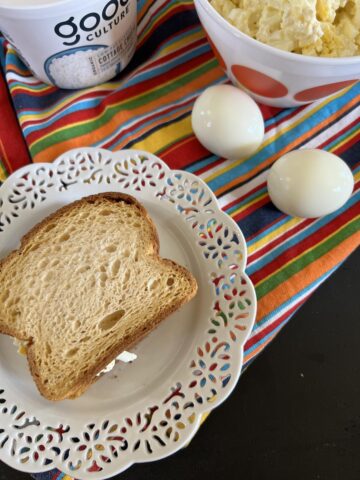 egg salad sandwich on whit e plate with two boiled eggs and a bowl of egg salad in background