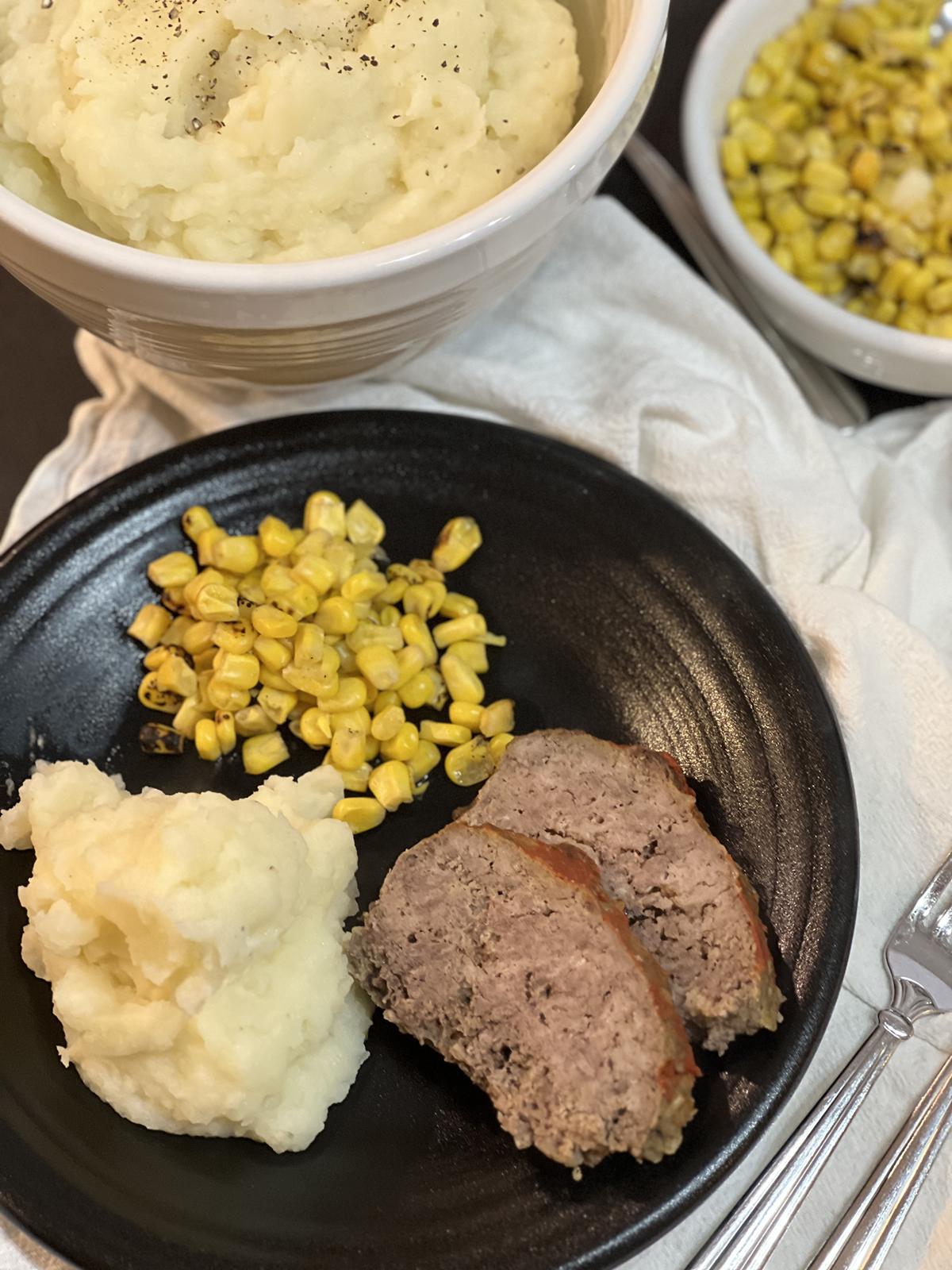 mashed potatoes, meatloaf, and corn on black plate with a white bowl full of mashed potatoes