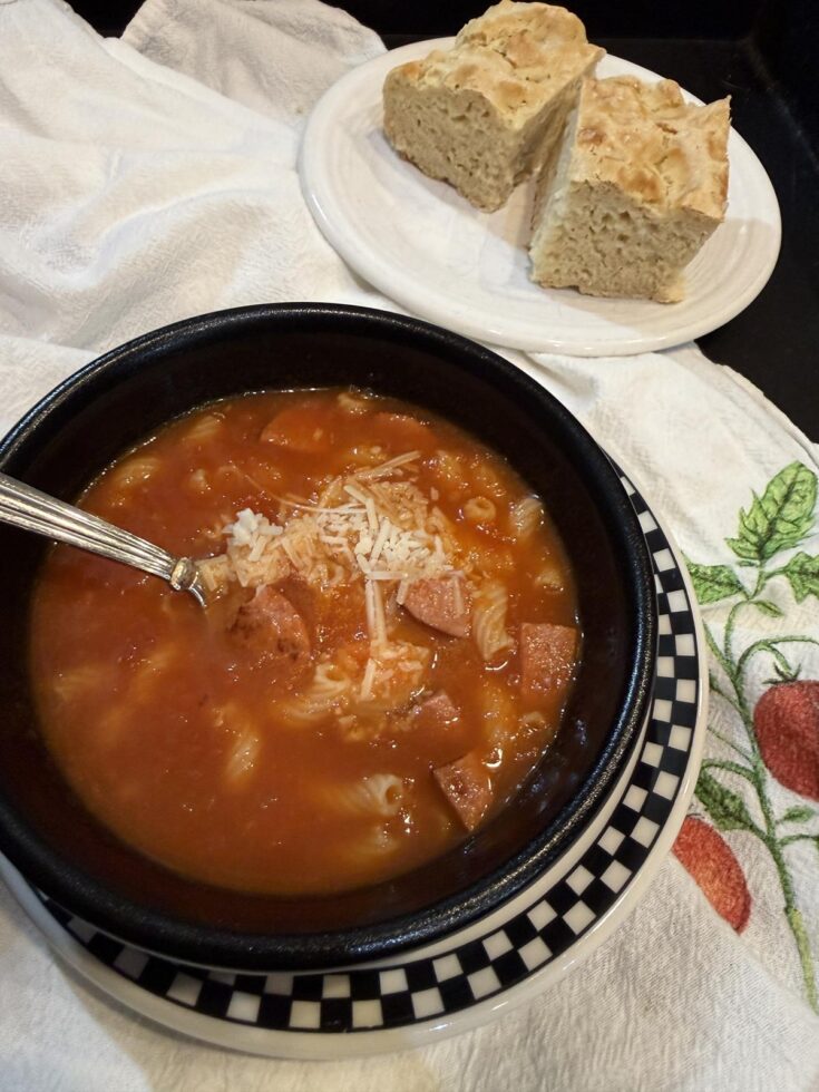 tomato soup with sausage and pasta in black bowl with spoon and cheese. Bread in background