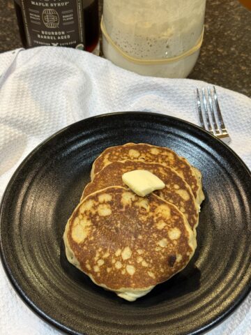 gluten free sourdough pancakes with butter on black plate on white napkin with sourdough starter in background