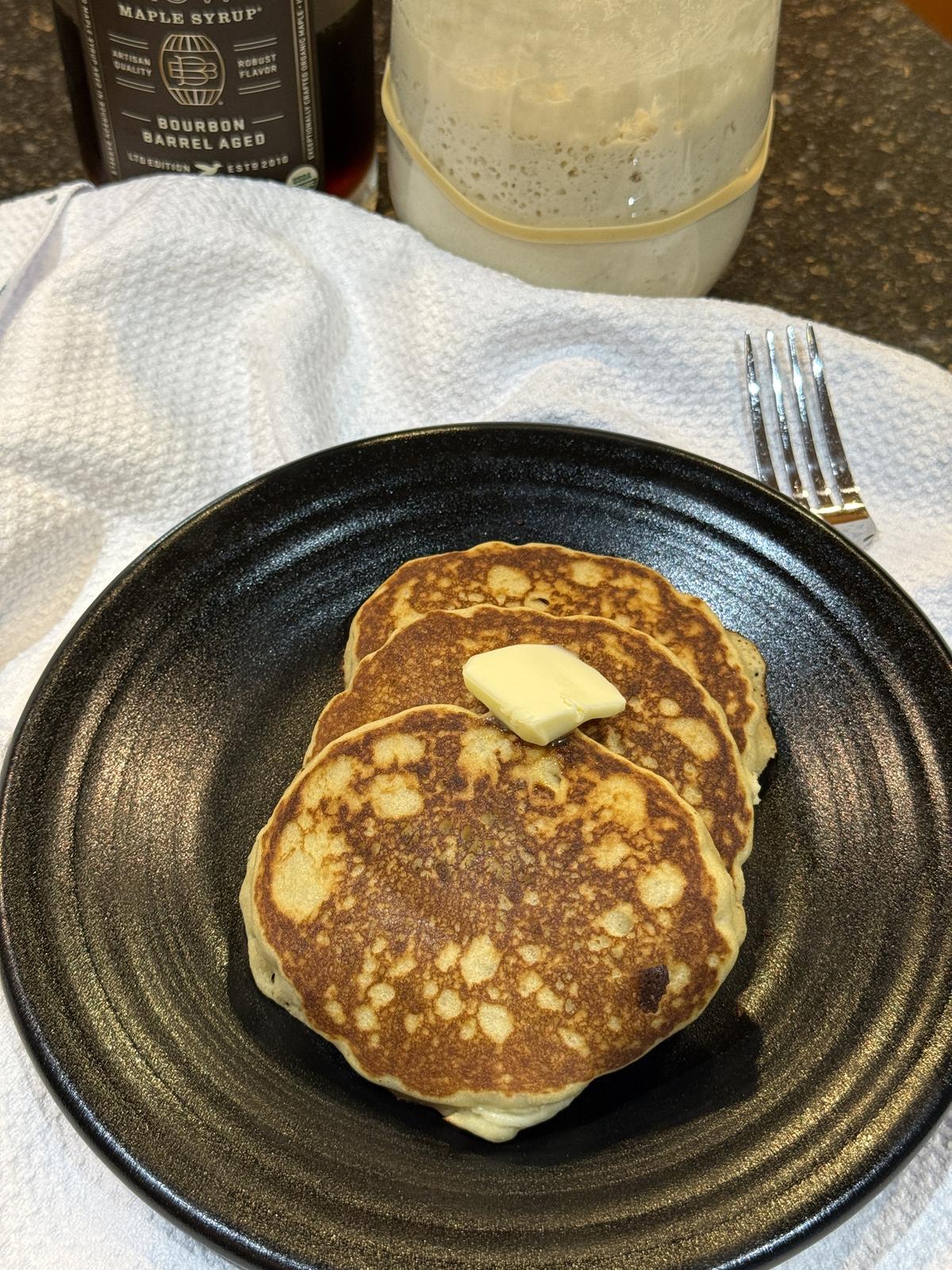 gluten free sourdough pancakes with butter on black plate on white napkin with sourdough starter in background
