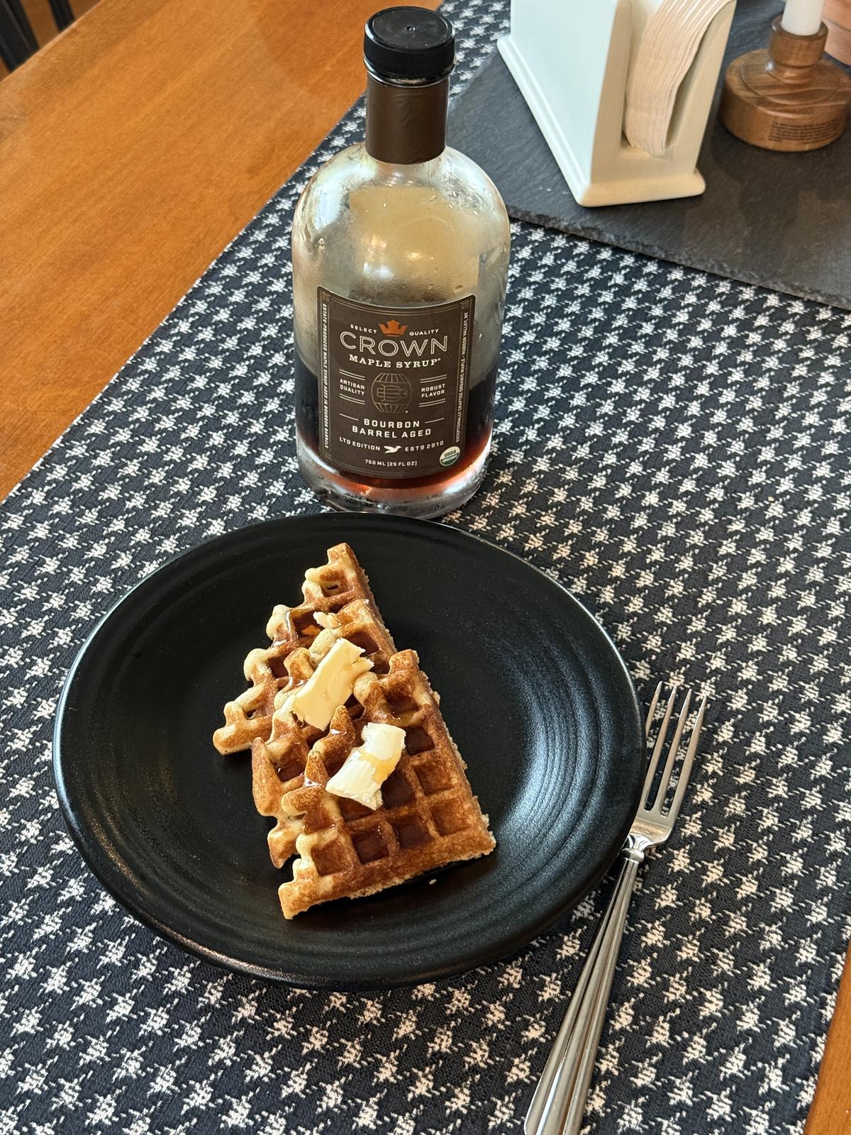 sourdough gluten free waffles with butter and syrup with a fork on a black plate with syrup bottle in background