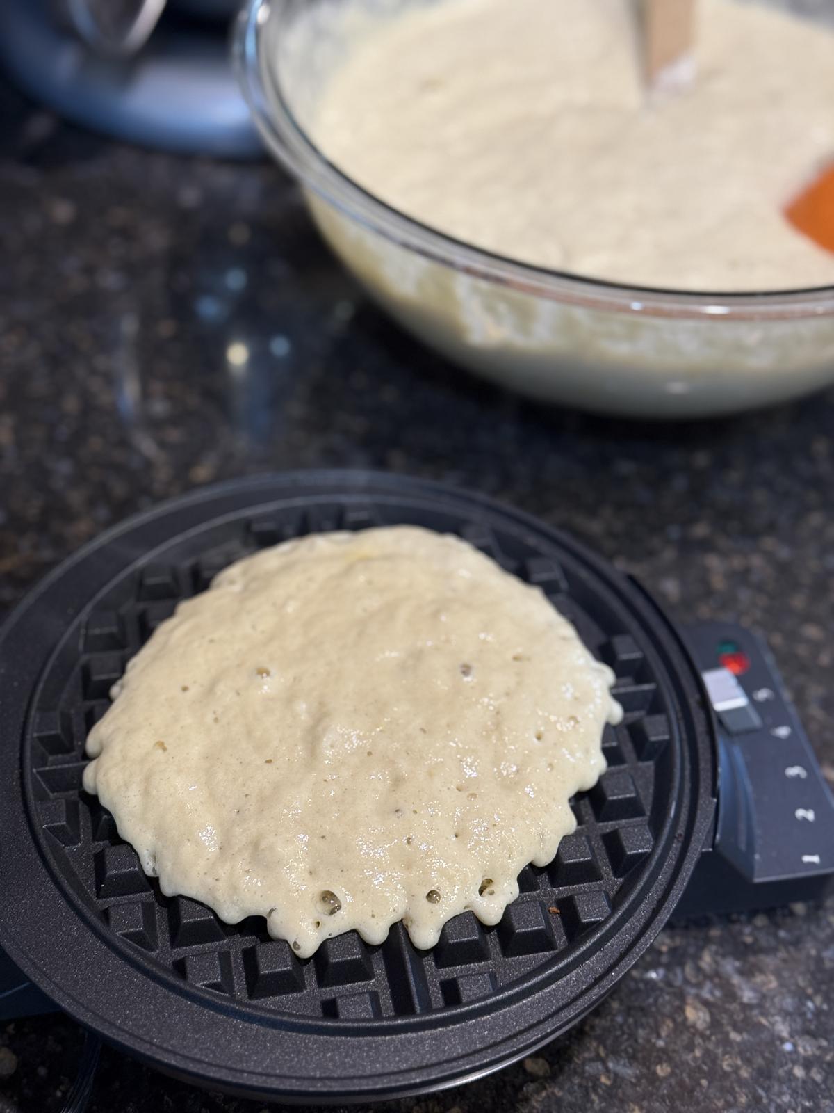 gluten free sourdough waffles cooking on a waffle iron with a bowl of waffle batter in background