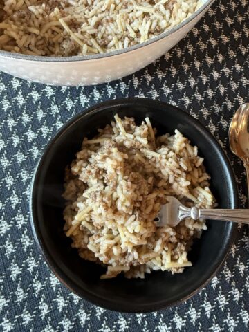 Homemade Rice A Roni with Ground Beef in black bowl with fork and white pan full of Rice A Roni on black and white table cloth