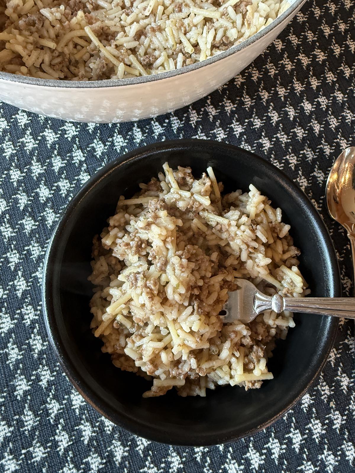 Homemade Rice A Roni with Ground Beef in black bowl with fork and white pan full of Rice A Roni on black and white table cloth