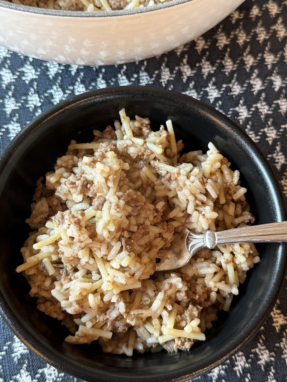 Homemade Rice A Roni with Ground Beef in black bowl with fork and white pan full of Rice A Roni on black and white table cloth